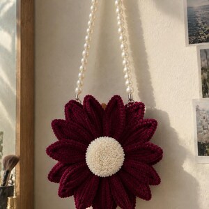 May include: A circular bag shaped like a flower with deep burgundy velvet-textured petals and a central white tufted disc. The bag features a long handle made of white faux pearls, allowing it to be hung from a circular gold-toned wall hook.