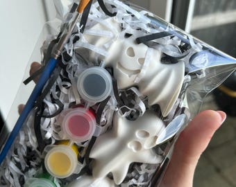 May include: A clear plastic bag containing three white ceramic Halloween-themed figures, including a ghost and two jack-o'-lanterns, nestled in black and white shredded paper. A blue-handled paintbrush and four small paint pots are included.