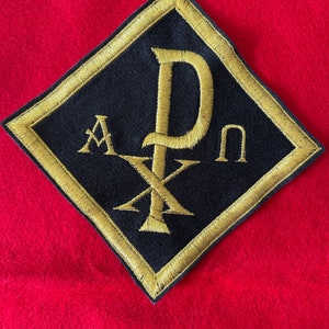 May include: A black diamond-shaped patch with a gold embroidered cross and the letters "A" and "P" on either side of the cross. The letters "D" and "n" are above and below the cross, respectively.