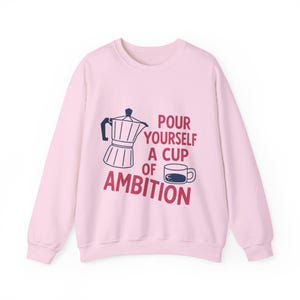 Puede incluir: Una sudadera de manga larga en color rosa claro con una ilustración en azul oscuro de una cafetera italiana y una pequeña taza de café. El texto impreso en rojo oscuro dice "POUR YOURSELF A CUP OF AMBITION" alrededor de los gráficos.