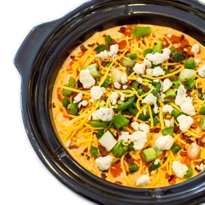 May include: A black ceramic slow cooker bowl filled with a creamy orange dip topped with shredded yellow cheddar cheese, crumbled white blue cheese, sliced green jalapeño peppers, chopped green onions, and small bits of red bacon.