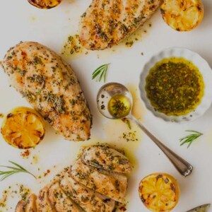 May include: Grilled chicken breasts seasoned with green herbs and oil, accompanied by halved charred lemons and a small white scalloped bowl filled with a green herb sauce, alongside a silver spoon containing a portion of the sauce.