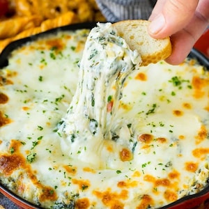 May include: A round red skillet filled with hot spinach and artichoke dip topped with melted white cheese and green herbs. A slice of toasted baguette is dipped into the center, pulling up long, stretchy strands of melted cheese from the dip.