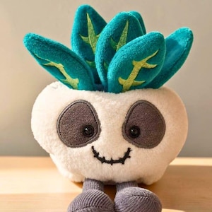 Puede incluir: Un juguete de peluche con forma de maceta blanca con una cara sonriente y ojos ovalados de color gris oscuro. La maceta contiene varias hojas de color verde azulado con detalles de nervaduras verde claro. Dos piernas de pana gris sobresalen.