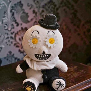 May include: A plush doll featuring a round white head with daisy flower eyes, a small black top hat, and a wide grinning mouth. The body is split into black and white halves with a ruffled collar, featuring a sun icon on one foot and a palm tree on the other.