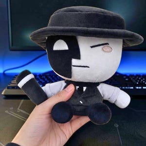 May include: A plush toy featuring a character with a split black and white face, wearing a black hat and a black suit with a white shirt. The character has one large white eye and one closed eye, with a simple line for a mouth and a small pink cheek.