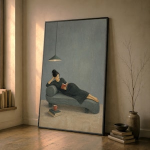 Puede incluir: Una impresión de arte mural vertical enmarcada que muestra a una persona de cabello oscuro con un vestido largo oscuro, recostada en una chaise longue gris mientras lee un libro rojo. Una lámpara colgante cuelga arriba y hay libros y una taza roja.