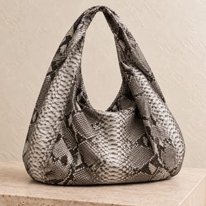 Könnte beinhalten: Eine Hobo-Handtasche mit einem Schlangenledermuster in Grau, Schwarz und Weiß. Die Tasche hat eine weiche, lockere Silhouette mit einem einzelnen integrierten Griff, der eine dreieckige Form bildet und eine detaillierte Schuppenstruktur zeigt.