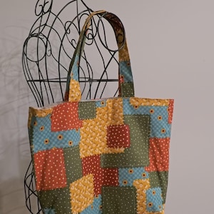 May include: A fabric tote bag featuring a patchwork design with square patterns in shades of olive green, mustard yellow, light blue, and terracotta orange. The bag has two matching fabric handles and a rectangular shape with visible stitching.