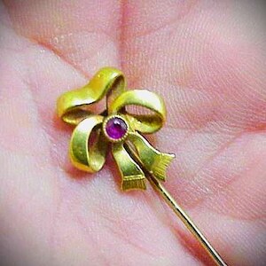 May include: A gold-colored bow pin with a small, round, red gemstone in the center. The pin is held in a hand, and the bow design has detailed curves and folds. The pin is a classic jewelry item.