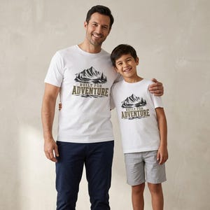 May include: A white short-sleeved t-shirt featuring a black and olive green graphic of a mountain range and pine trees with the text "BUILT FOR ADVENTURE" printed in a bold, blocky font centred on the chest area of the garment.