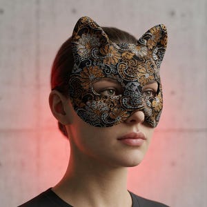 Pode incluir: Uma máscara em forma de gato com uma base preta e um padrão floral e paisley intrincado em tons metálicos de ouro e prata. A máscara cobre a parte superior do rosto, incluindo a área dos olhos, e tem duas orelhas pontiagudas no topo.