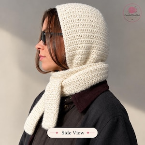 May include: A cream-colored crochet hood with an integrated scarf, featuring a dense, uniform stitch pattern. The hood covers the head and wraps around the neck, with two long ends hanging down. The design is simple, textured, and solid-colored.