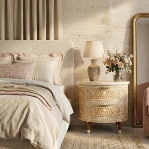 Puede incluir: Una mesita de noche redonda de dos cajones con un intrincado patrón floral incrustado de color crema sobre un acabado de madera clara, sostenida por cuatro patas cónicas. Una lámpara de mesa beige y un florero de cristal con rosas rosa pálido.
