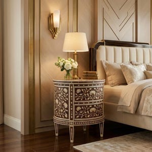 May include: A round wooden nightstand featuring two drawers with intricate white floral inlay patterns. The piece stands on four tapered legs and is topped with a table lamp, a small glass vase containing white flowers, and three stacked books.