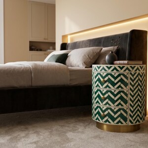 May include: A cylindrical three-drawer nightstand featuring a green and white chevron pattern inlay, three small round brass knobs, and a circular brass base. The piece is positioned next to a bed with a dark headboard and neutral bedding.