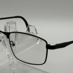 Op de afbeelding: Een bril met een dun zwart metalen montuur en heldere glazen. De bril heeft rechthoekige randen, slanke pootjes en verstelbare neuspads, rustend op een doorzichtige acrylstandaard om de volledige structuur van de bril te tonen.