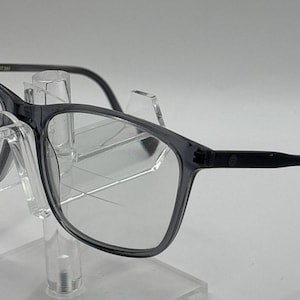 May include: A pair of translucent dark gray eyeglasses with a rectangular frame shape. The lenses feature visible horizontal lines indicating a multifocal or progressive lens design. The temples are thin and dark gray, matching the frame front.