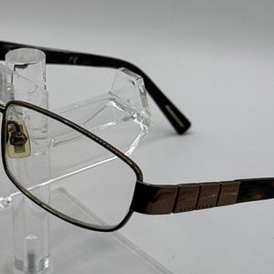 May include: A pair of eyeglasses with a metallic brown rectangular frame. The temples feature a textured brown metal section transitioning into a dark tortoiseshell pattern at the tips. The clear lenses are held by a thin, durable metal rim.