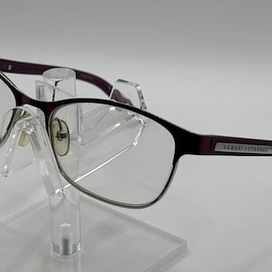 May include: Dark purple eyeglasses with clear lenses and a silver-toned metal frame. The temples feature a rectangular silver plate with the text Armani Exchange engraved in black. The glasses have adjustable nose pads and thin, dark purple arms.