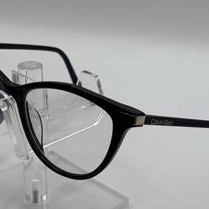 Może przedstawiać: Czarne okulary typu kocie oczy z przezroczystymi szkłami i cienkimi czarnymi zausznikami. Nazwa marki Calvin Klein jest nadrukowana złotymi literami na zewnętrznej stronie lewego zausznika. Oprawki mają błyszczące wykończenie i srebrne metalowe akcenty.