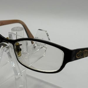 May include: Black eyeglasses with clear lenses and a light pink interior frame. The temples feature a gold-colored decorative pattern near the hinges. The frames are constructed with a dark exterior and a contrasting light pink inner surface.