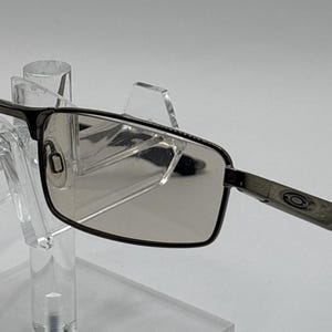 May include: Rectangular eyeglasses with a dark metallic frame and tinted lenses. The temples feature a distinct logo detail near the hinges and black tips. These glasses have adjustable nose pads and a sleek, structured design for clear vision.
