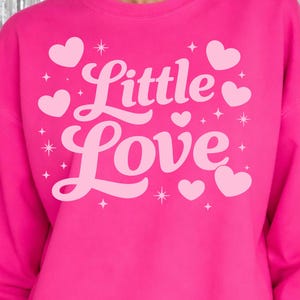 May include: A bright pink sweatshirt with the words "Little Love" in a cursive font, surrounded by small hearts and stars. The sweatshirt is a solid color and has a crew neck.