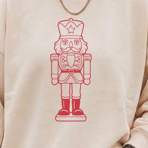 May include: A cream-colored sweatshirt featuring a red outline of a nutcracker soldier. The nutcracker design includes a star-topped hat, a detailed face, and a uniform with boots. The sweatshirt has long sleeves and a crew neck.