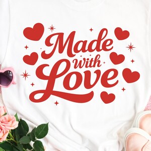 Könnte beinhalten: Weißes T-Shirt mit dem Schriftzug "Made with Love" in roter Schrift und Herzmotiven. Rosa herzförmige Sonnenbrille, rosa Turnschuhe und ein Strauß rosa Rosen sind um das Shirt herum auf einer rosa-weiß karierten Oberfläche angeordnet.