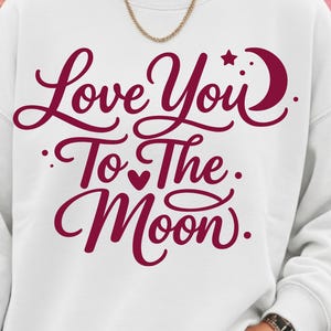 May include: White sweatshirt with the phrase "Love You To The Moon" in burgundy script. The design includes a crescent moon and star. The sweatshirt is a casual style, suitable for everyday wear.