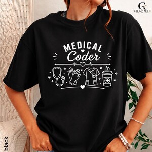Op de afbeelding: Een zwart T-shirt met korte mouwen en witte grafische tekst met de tekst MEDICAL Coder. Het ontwerp bevat iconen van een stethoscoop, medische handschoenen, een medisch shirt en een koffiebeker met een kruissymbool, geaccentueerd door hartjes.
