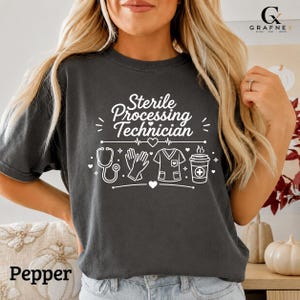 Könnte beinhalten: Ein anthrazitfarbenes Kurzarm-T-Shirt mit weißem Aufdruck Sterile Processing Technician. Das Design enthält grafische Symbole eines Stethoskops, medizinischer Handschuhe, eines Kasacks, eines Kaffeebechers und kleiner Herzen.