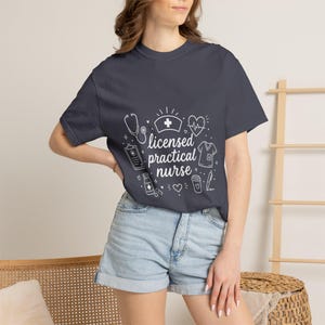 Könnte beinhalten: Ein dunkelgraues Kurzarm-T-Shirt mit weißen Strichzeichnungen medizinischer Symbole wie Stethoskop, Krankenschwesterhaube, Herz, Klemmbrett und Pillendose, sowie dem mittig aufgedruckten Text "licensed practical nurse".