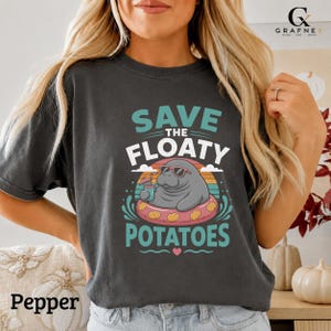 May include: A charcoal gray t-shirt featuring a graphic of a manatee wearing sunglasses and floating in a pink inner tube while holding a drink. The text reads SAVE THE FLOATY POTATOES in teal and white, set against a sunset background.