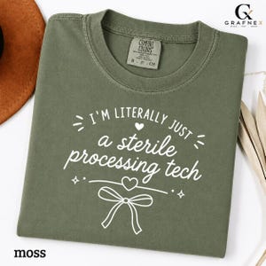 Könnte beinhalten: Ein moosgrünes Kurzarm-T-Shirt mit weißem Text I'M LITERALLY JUST a sterile processing tech, ergänzt durch ein kleines Herz und eine dekorative Schleifengrafik. Das T-Shirt hat einen Rundhalsausschnitt und ein Comfort Colors Markenetikett.