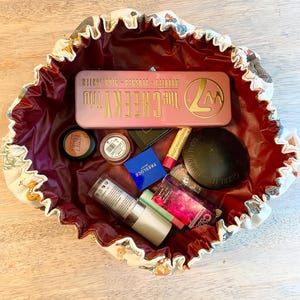 May include: A round, drawstring makeup bag with a cream-colored exterior featuring a multi-colored pattern and a deep burgundy interior. The bag is filled with various cosmetics, including a pink W7 The Cheeky Trio palette, a black Guerlain compact, a blue Tresluce Beauty box, a cylindrical bottle, and several small makeup containers.