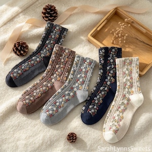 May include: Five pairs of crew-length socks featuring a cable knit texture and a colourful floral pattern. The socks are displayed in charcoal, brown, light grey, navy blue, and cream colours, each with a solid toe and heel section.