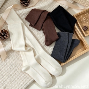 Puede incluir: Una colección de cuatro pares de calcetines en colores sólidos que incluyen crema, marrón oscuro, negro y gris carbón. Los calcetines están dispuestos en una composición plana, con una textura de punto suave y un diseño estándar hasta el tobillo.