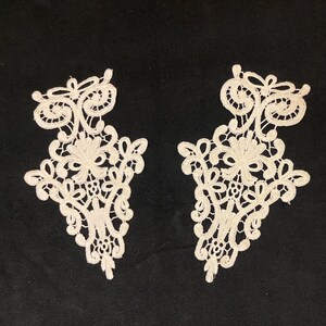 May include: Two off-white lace appliques with intricate floral and scrollwork designs. The appliques are symmetrical, with a teardrop shape and delicate, openwork patterns. They are set against a black background.
