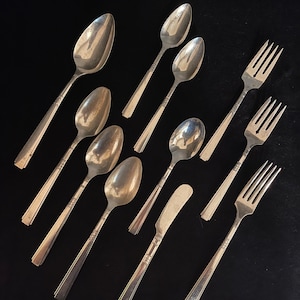 May include: A set of silverware, including spoons, forks, and a butter knife, all with a silver finish. The silverware is arranged on a black surface.
