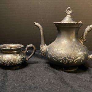 May include: Antique silver tea set, featuring a teapot with a decorative lid and a sugar bowl with two handles. Both pieces have ornate floral engravings. The set is displayed on a dark surface.