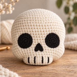 May include: A round, off-white crocheted skull with large, circular black eye sockets, a small triangular black nose, and four vertical black stitched lines representing teeth on the lower jaw, crafted with a visible yarn texture.