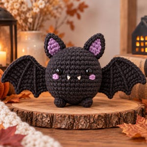 May include: A round, black crocheted bat plush toy featuring large black eyes, small white fangs, and light purple accents on the inner ears and cheeks. The bat has two outstretched wings and two small feet, crafted with a textured yarn stitch pattern.