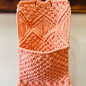 May include: A coral-colored macrame wall hanging featuring a woven pocket design suspended from a circular wooden ring. The piece is crafted with intricate knotted patterns and finished with a fringe at the bottom edge, creating a textured storage pouch.