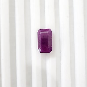 May include: A rectangular cut gemstone with a deep magenta and purple hue. The stone features faceted edges that catch the light, displaying a translucent quality with visible internal inclusions throughout the body of the polished crystal.