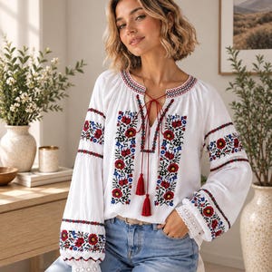 Puede incluir: Una blusa blanca de manga larga con intrincados bordados florales en rojo, azul y negro en la parte delantera y las mangas. La prenda incluye un escote en V con finos cordones rojos que terminan en borlas y encaje blanco en los puños.