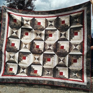 May include: A large quilt with a log cabin pattern in shades of brown, white, and red. The quilt is being held up by two people.