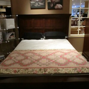 May include: A white bed with a pink and green quilt. The bed has a wooden headboard with a dark wood finish.