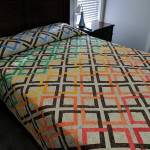 May include: A colorful quilt with a geometric pattern of squares and rectangles in shades of blue, green, yellow, orange, and red. The quilt is made with a white fabric base and has a brown border.